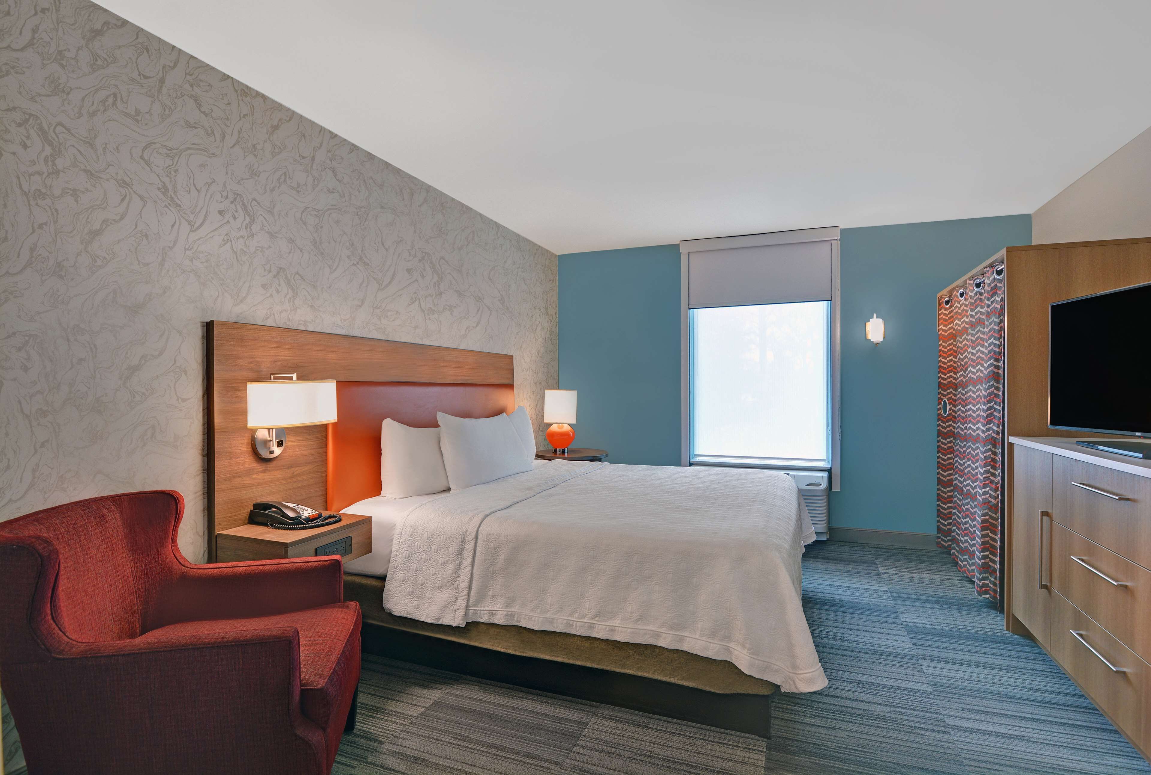 A hotel room with a bed, a red armchair, a TV, and a window.
