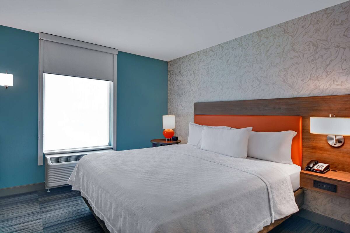 A hotel room with a large bed, a nightstand with a lamp, a telephone, and a window with blinds.