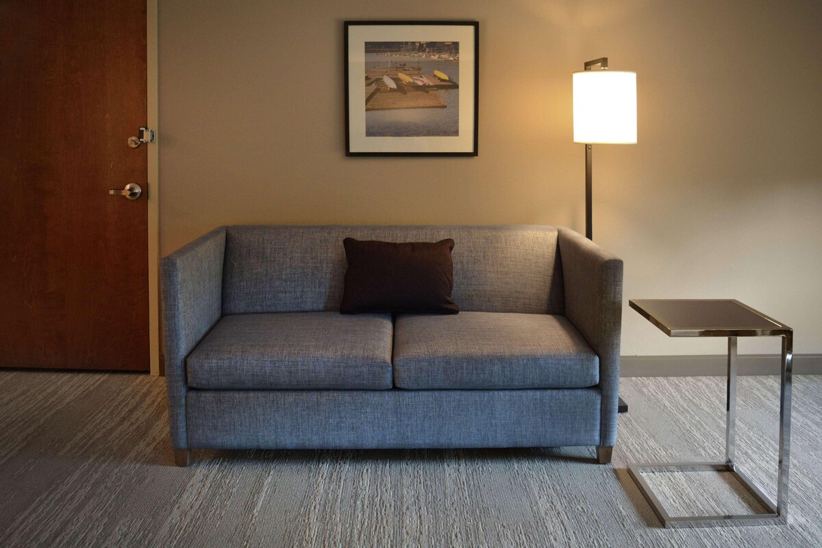 A room with a grey sofa, a framed picture on the wall, a lamp, and a small table.