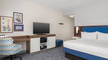 Quarto de hotel com cama, escrivaninha, televisão, cadeira e mesa pequena.