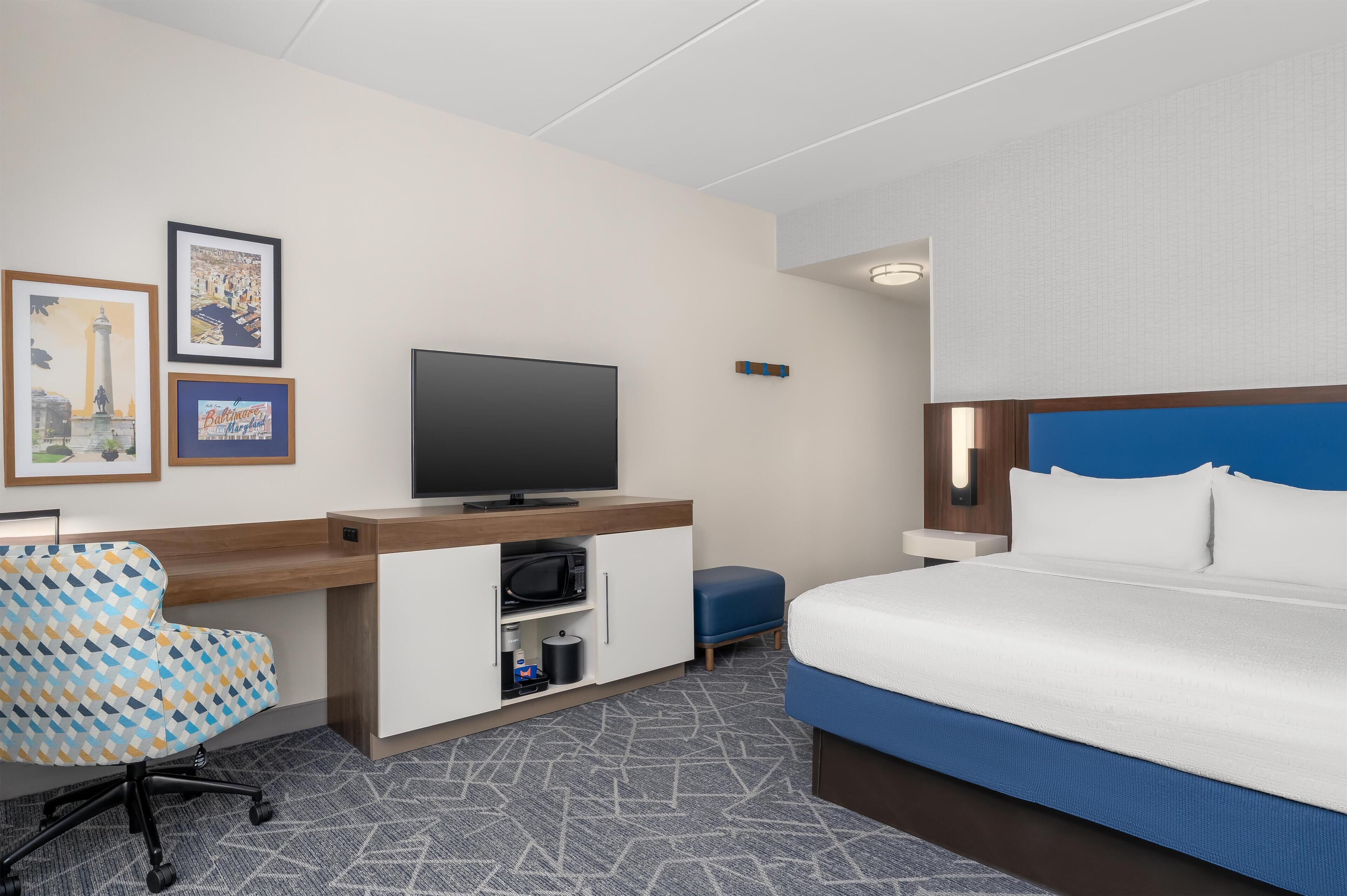 A hotel room with a bed, a desk with a television, a chair, and a small table.