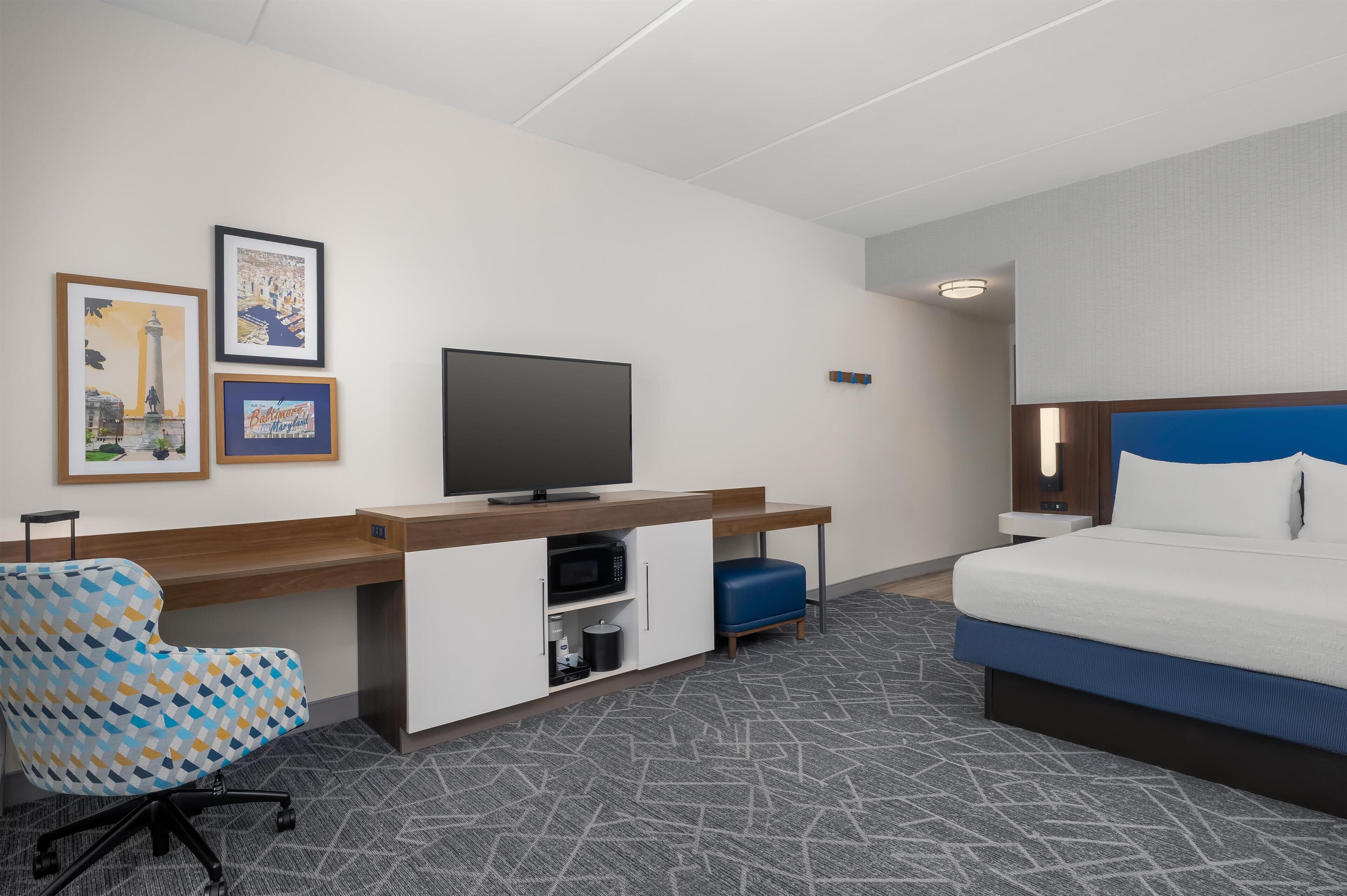 Una habitación de hotel con cama, un escritorio con televisor, una silla y dos cuadros en la pared.