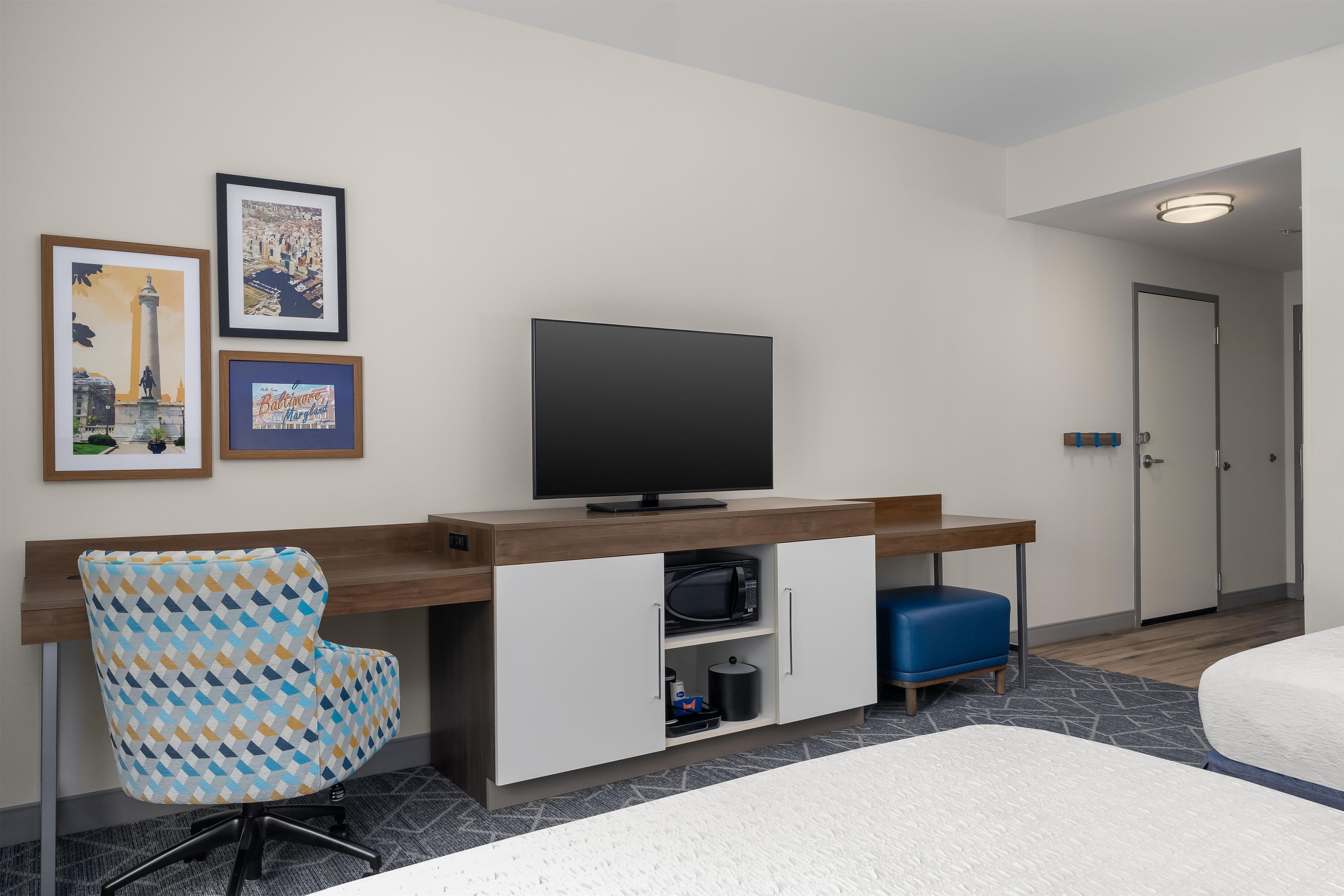 Habitación de hotel con cama, escritorio con televisor de pantalla plana, sillón estampado y mueble con microondas.