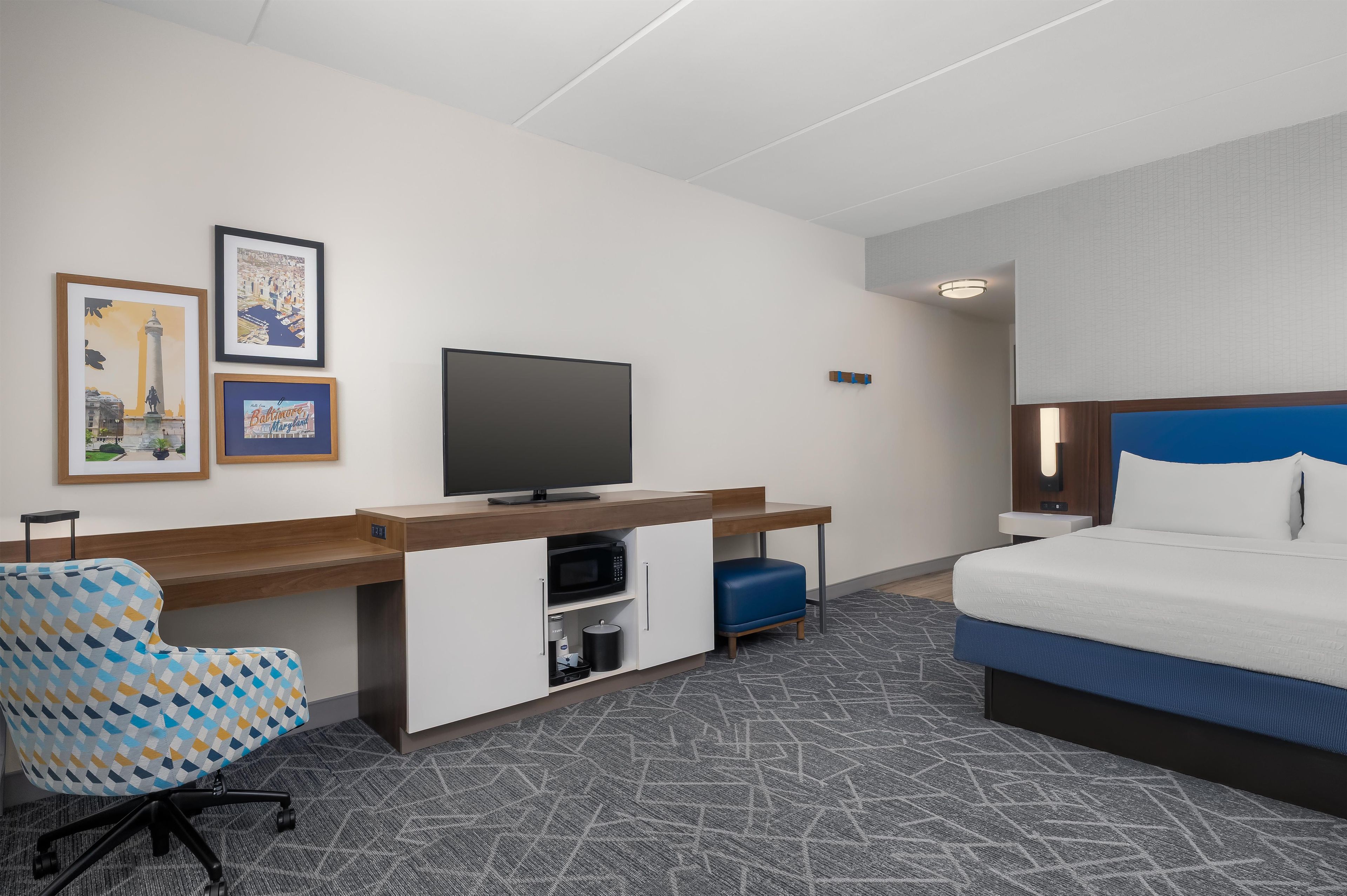 A hotel room with a bed, a desk with a television, a chair, and two framed pictures on the wall.