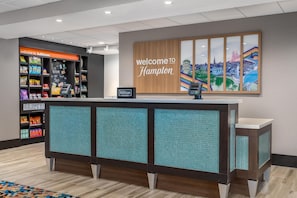 A reception desk with a 'Welcome to Hampton' sign, a snack bar with various packaged goods, and a cityscape artwork on the wall. - Hampton Inn Baltimore Bayview Campus (Baltimore)