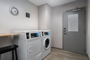 A laundry room with two front-loading washing machines, a clock, and a door. - Hampton Inn Baltimore Bayview Campus (Baltimore)