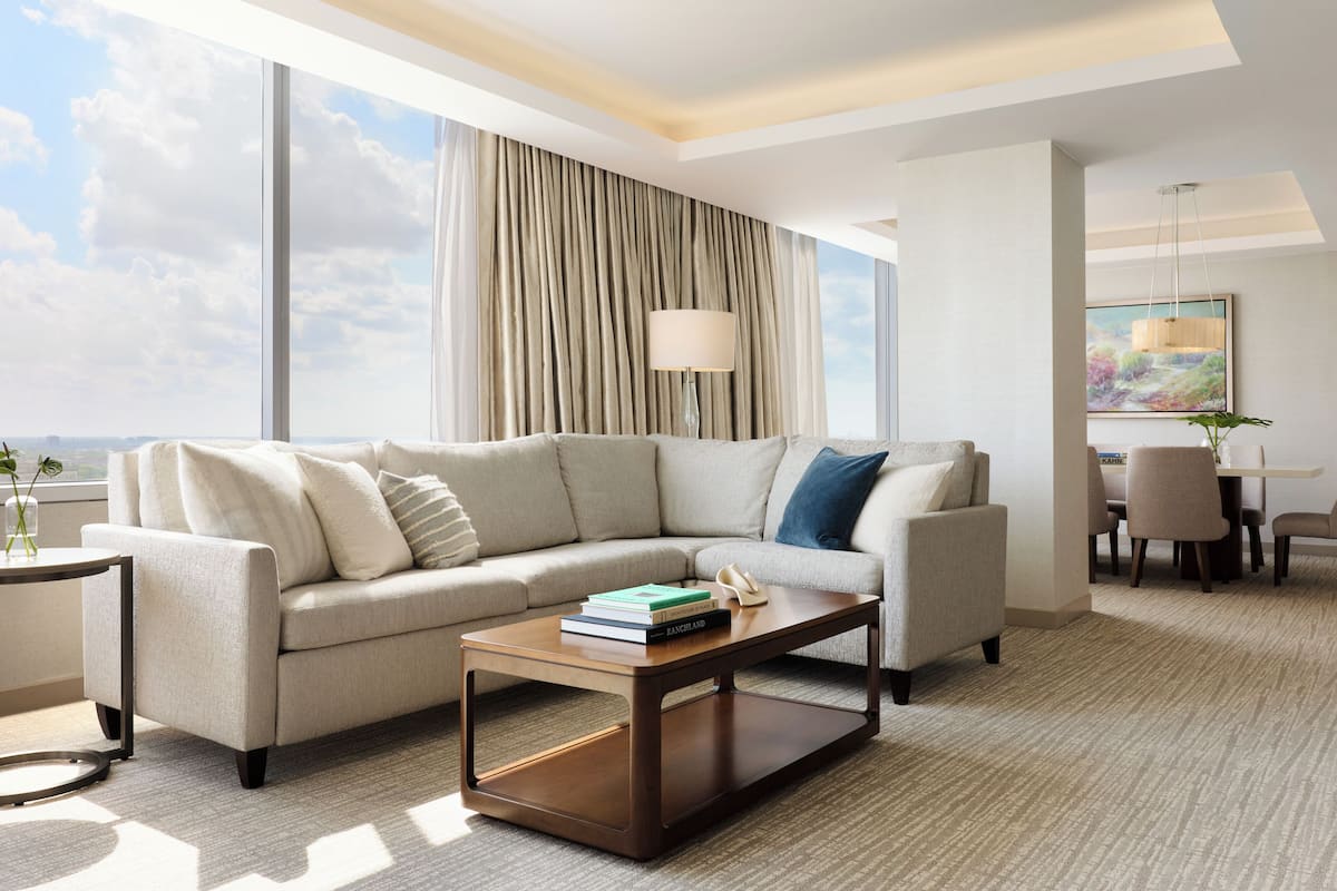 A modern hotel room with a large window, a sofa, a coffee table, and a dining area.