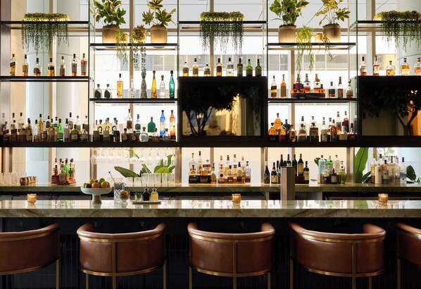 Un espace bar doté de plusieurs étagères remplies de bouteilles d’alcool, de plantes suspendues et de tabourets de bar.