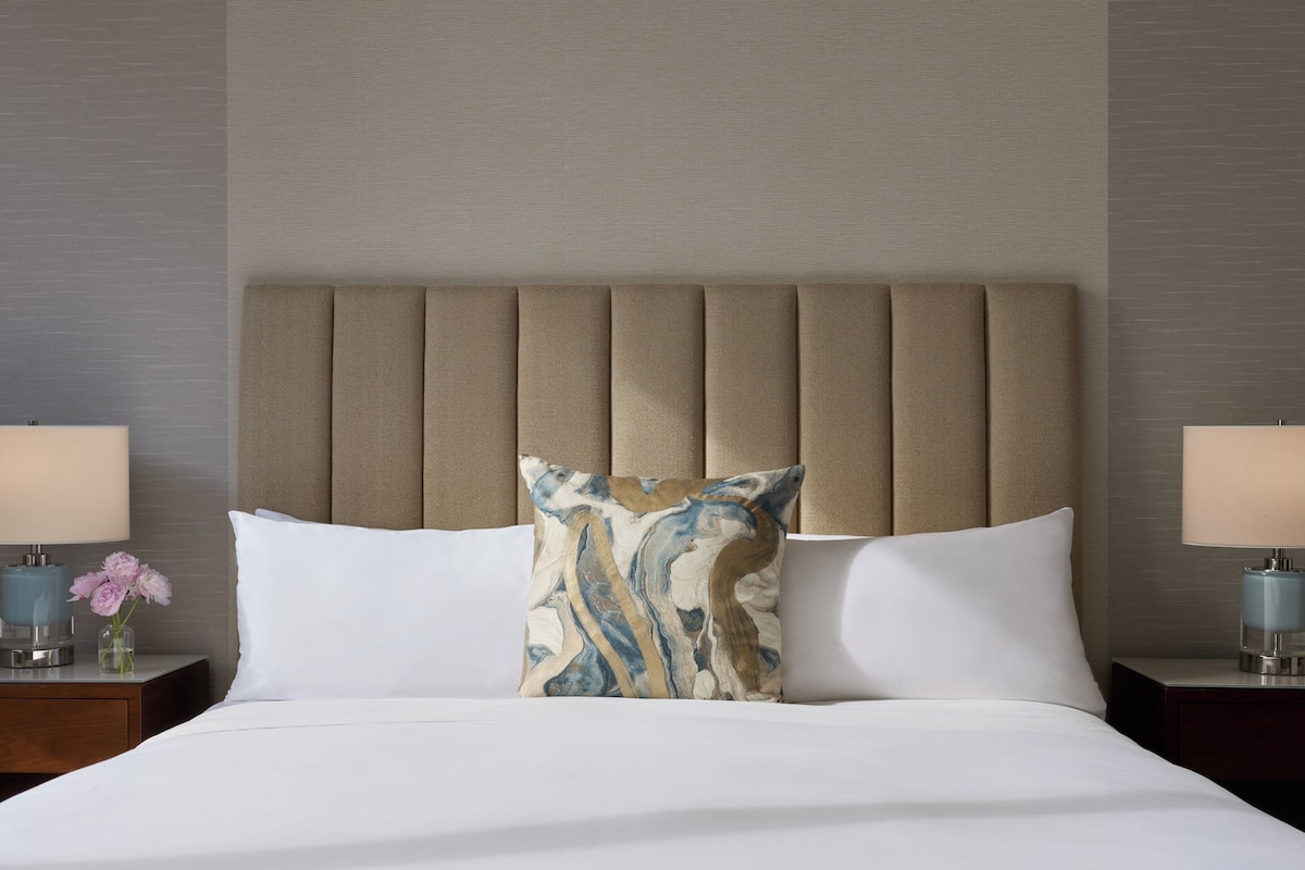 A hotel room with a bed, two nightstands, a beige upholstered headboard, and a decorative pillow.