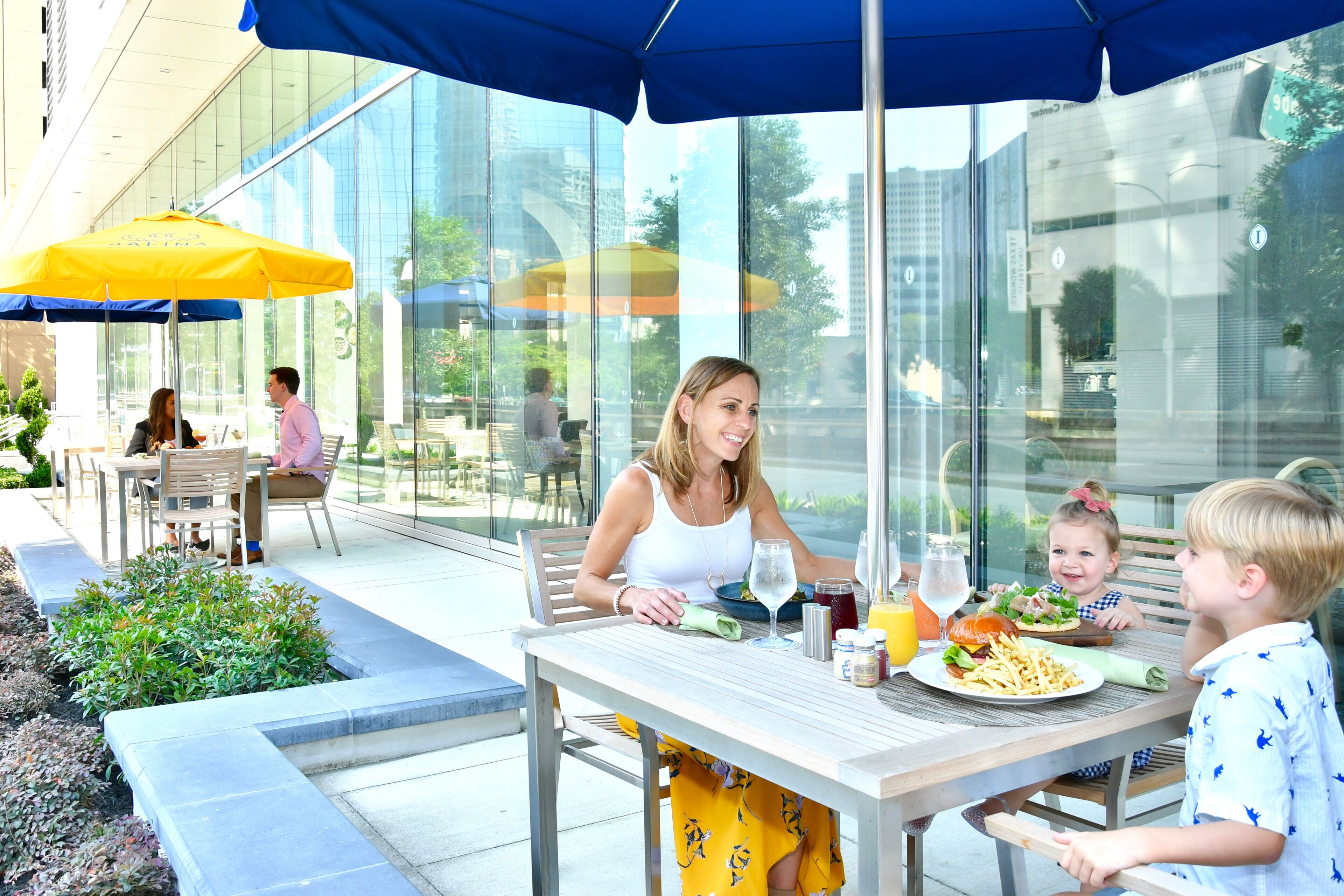 An outdoor dining area with tables and chairs, umbrellas, and a glass building in the background.