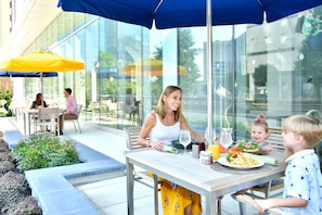 An outdoor dining area with tables and chairs, umbrellas, and a glass building in the background.