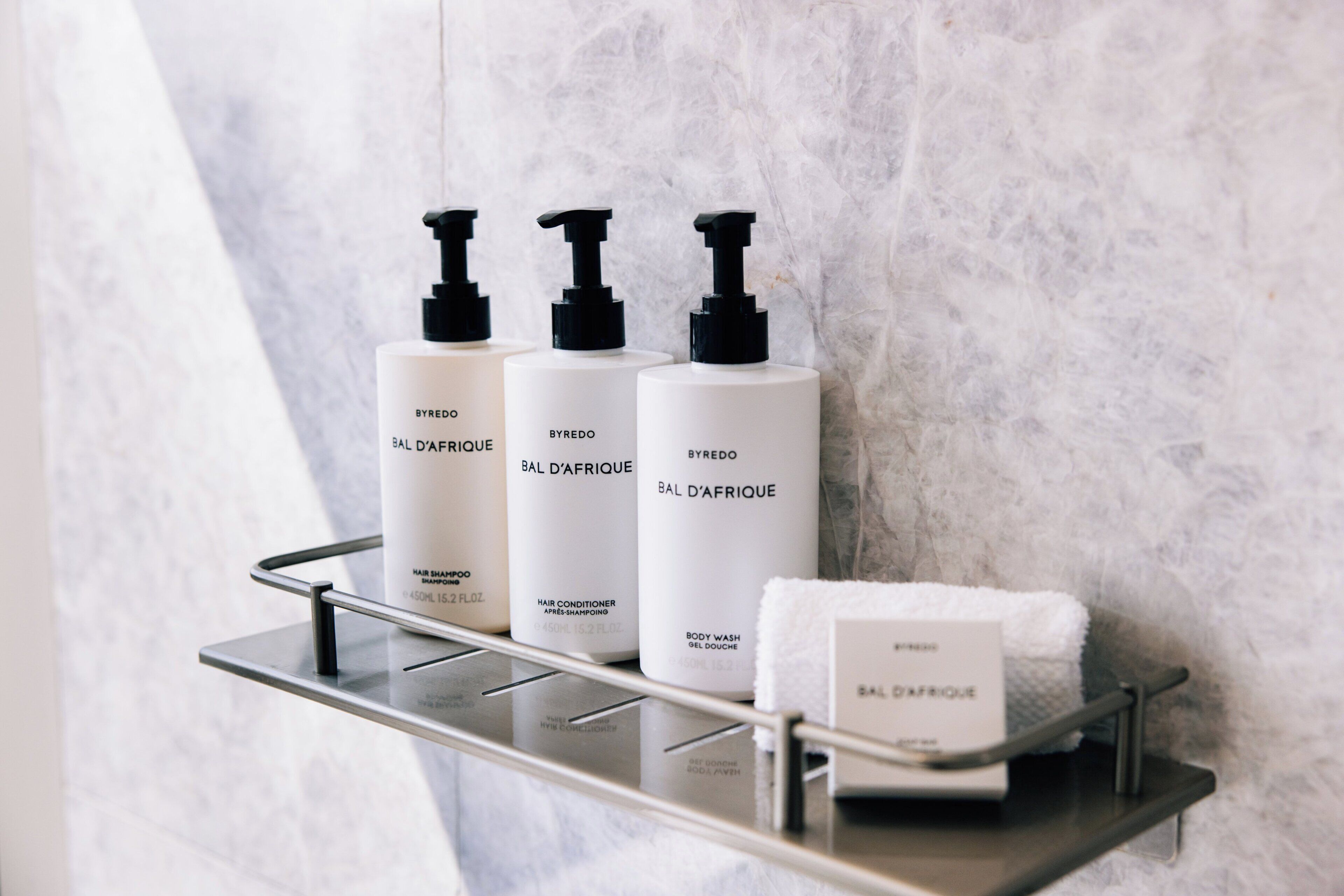 A bathroom shelf with three white bottles of BYREDO BAL D'AFRIQUE hair products and a white towel.