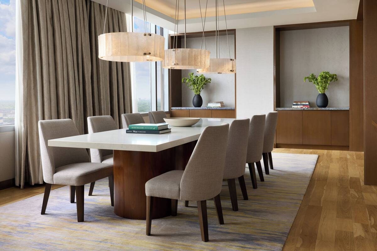 A modern dining area with a large table, multiple chairs, hanging pendant lights, and a view of the city through large windows.