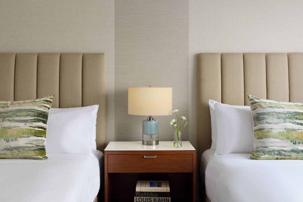 A hotel room with two beds, a nightstand with a lamp, a vase with flowers, and a book titled 'Architecture of Place' by Louis Kahn.