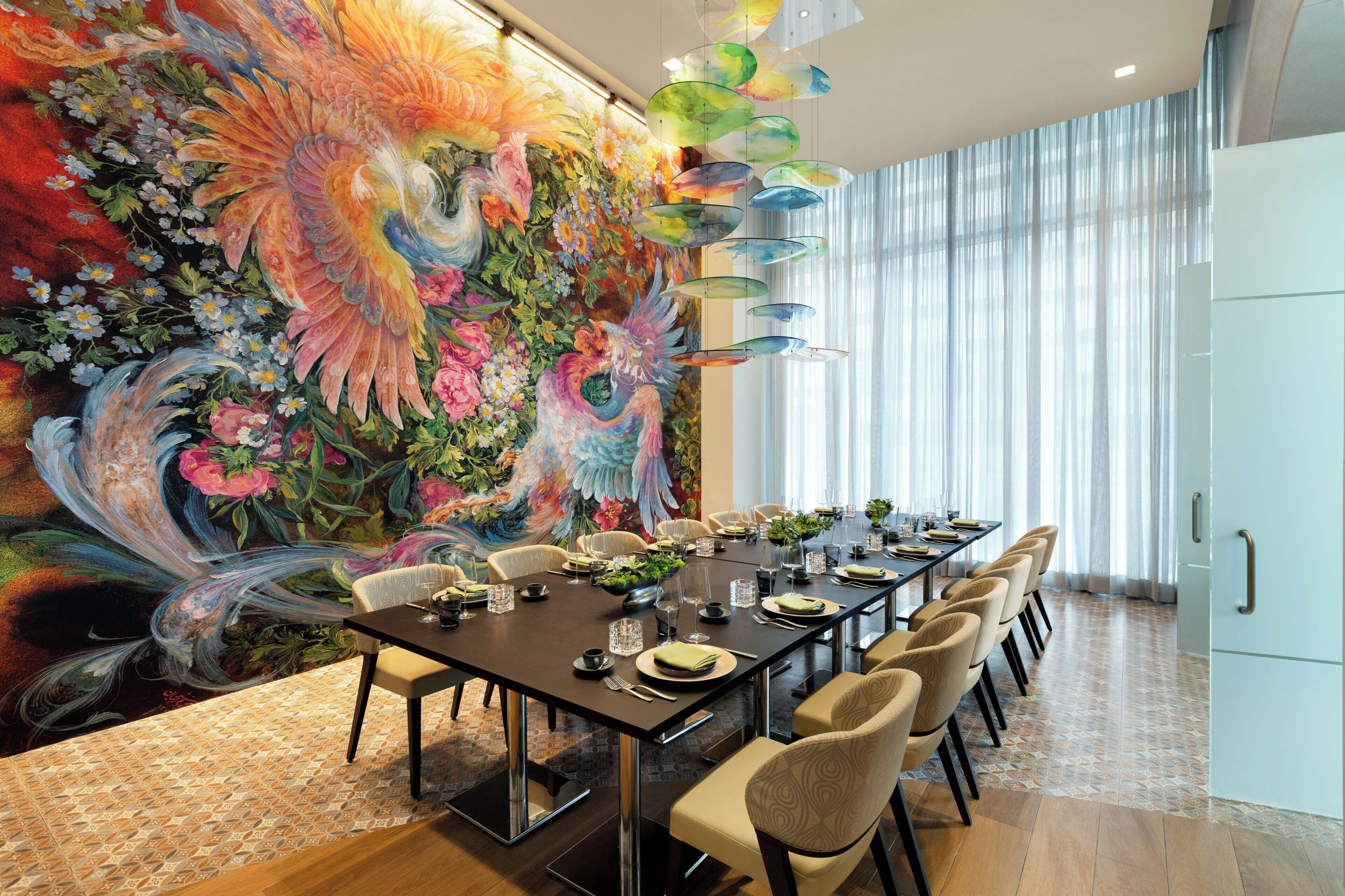 A dining area with a long table set for six, surrounded by chairs, with a large floral mural on the wall behind.