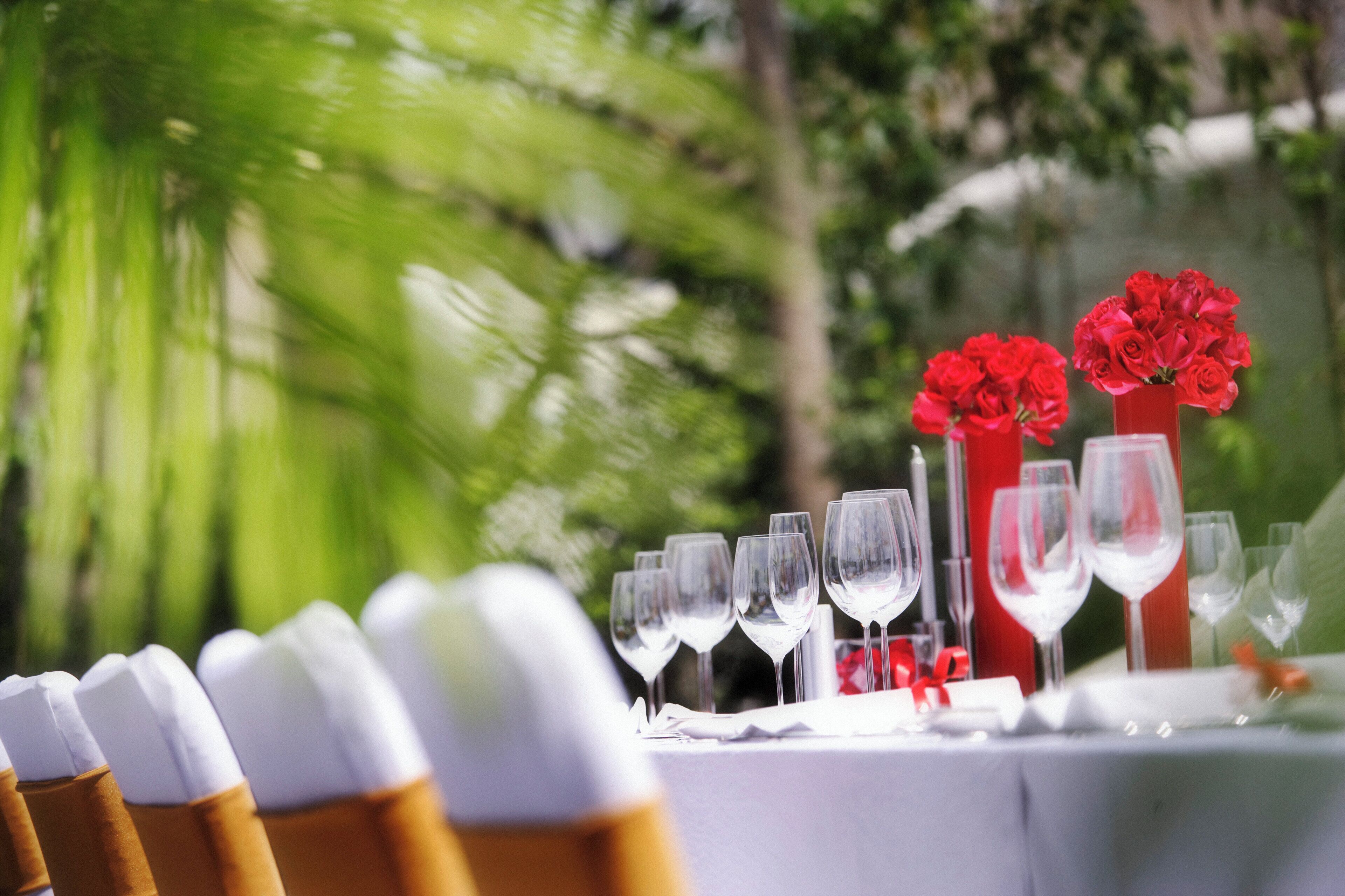 A table set with wine glasses and a red vase with flowers, outdoors with greenery in the background.