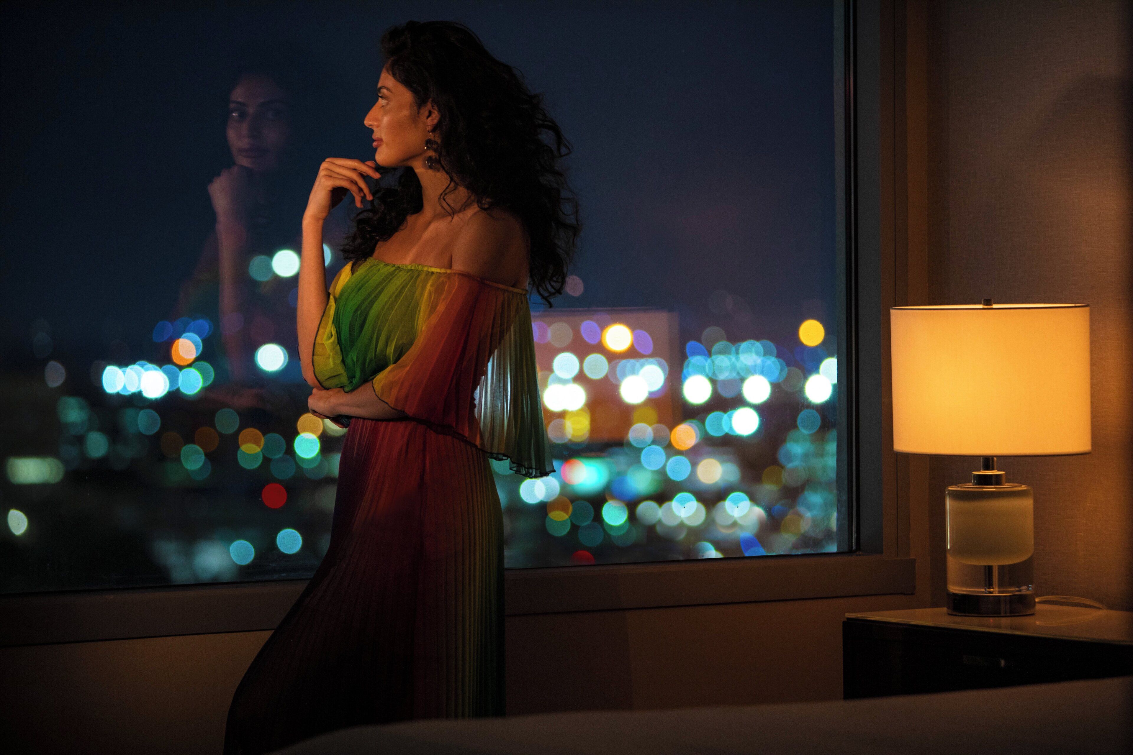 A person in a room with a view of a cityscape at night, a lamp on a bedside table, and a window.