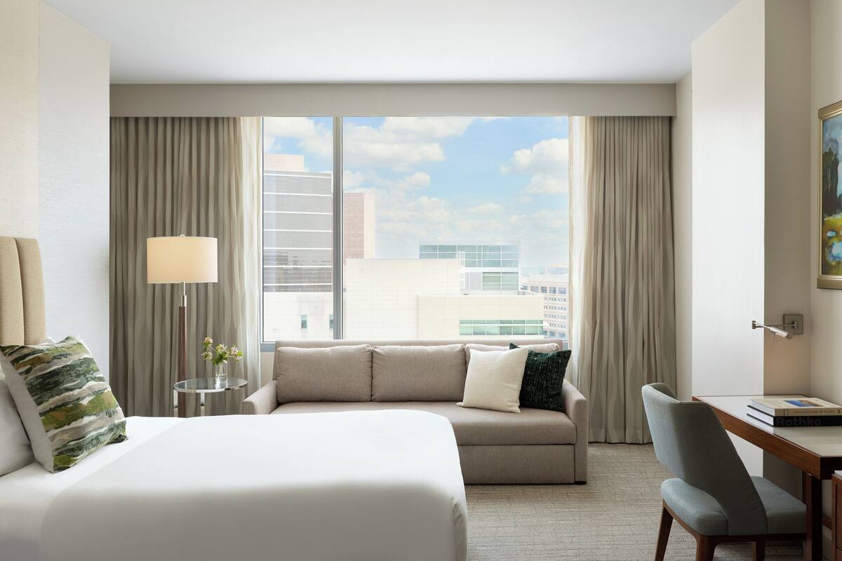 A hotel room with a bed, a sofa, a desk, a lamp, and a view of buildings through large windows.
