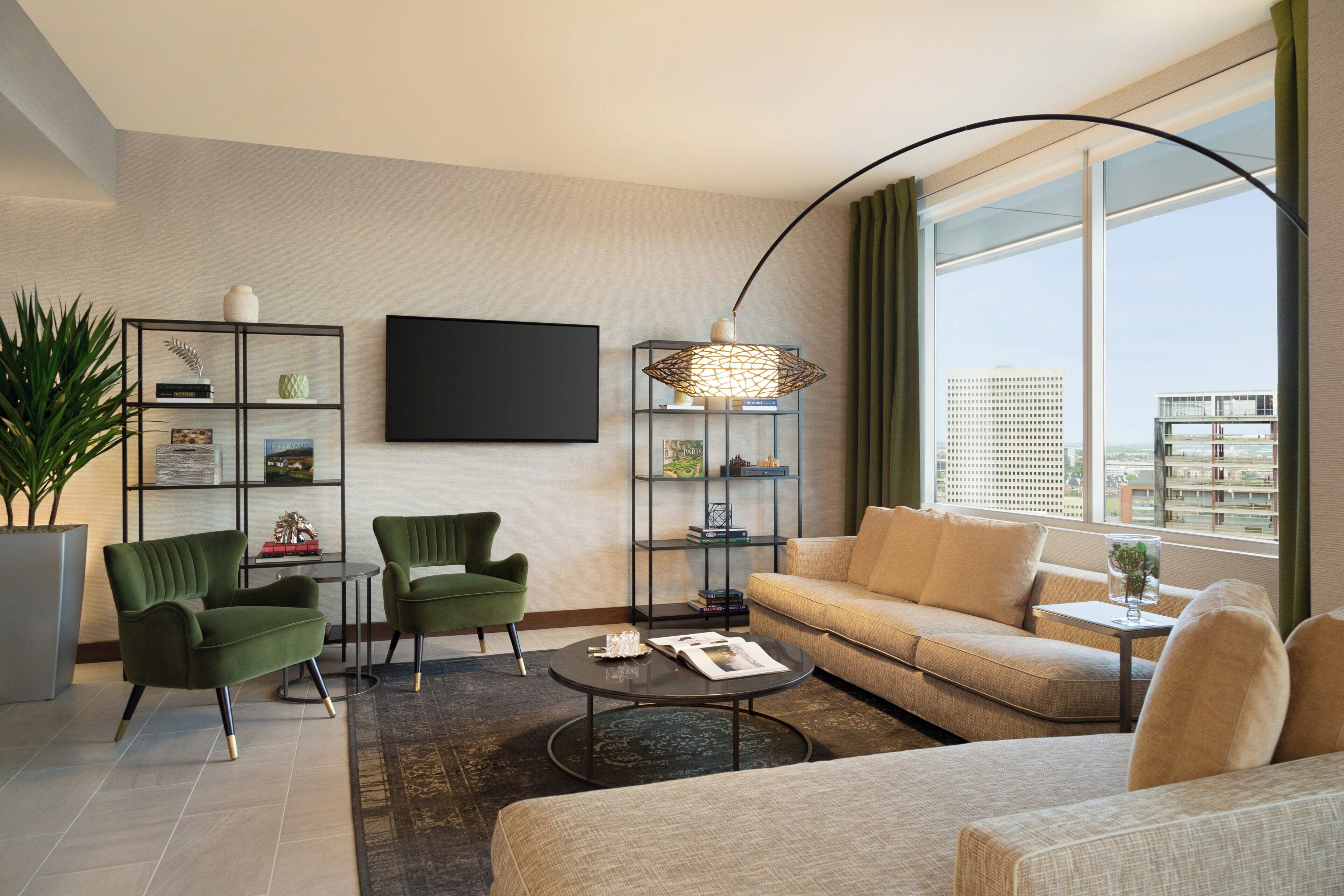 Uma sala de estar moderna com um sofá, duas poltronas, uma mesa de centro e uma TV. Há estantes com objetos decorativos e uma janela grande com vista para edifícios.