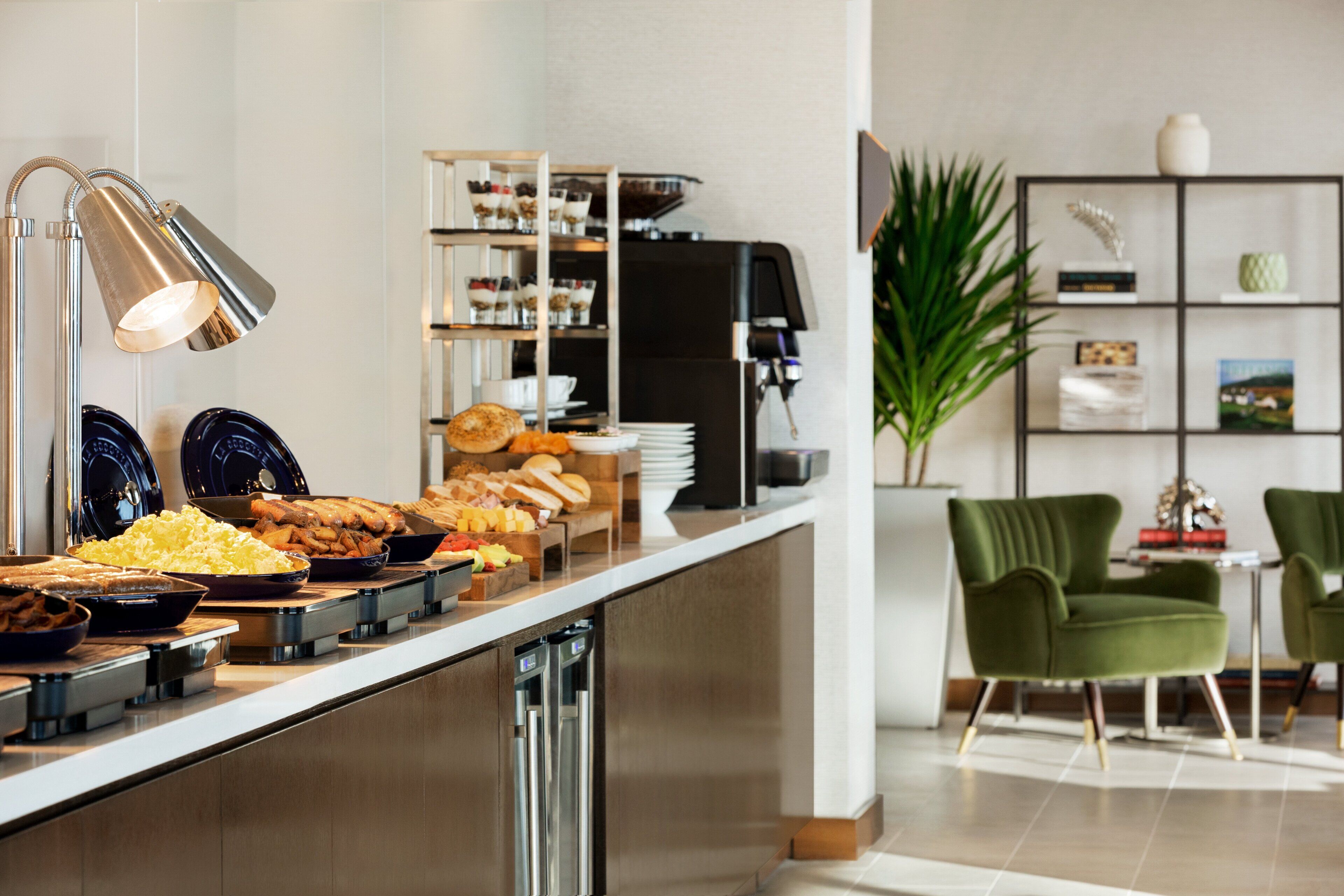 A buffet with various food items, a coffee machine, and a seating area with chairs and a plant.