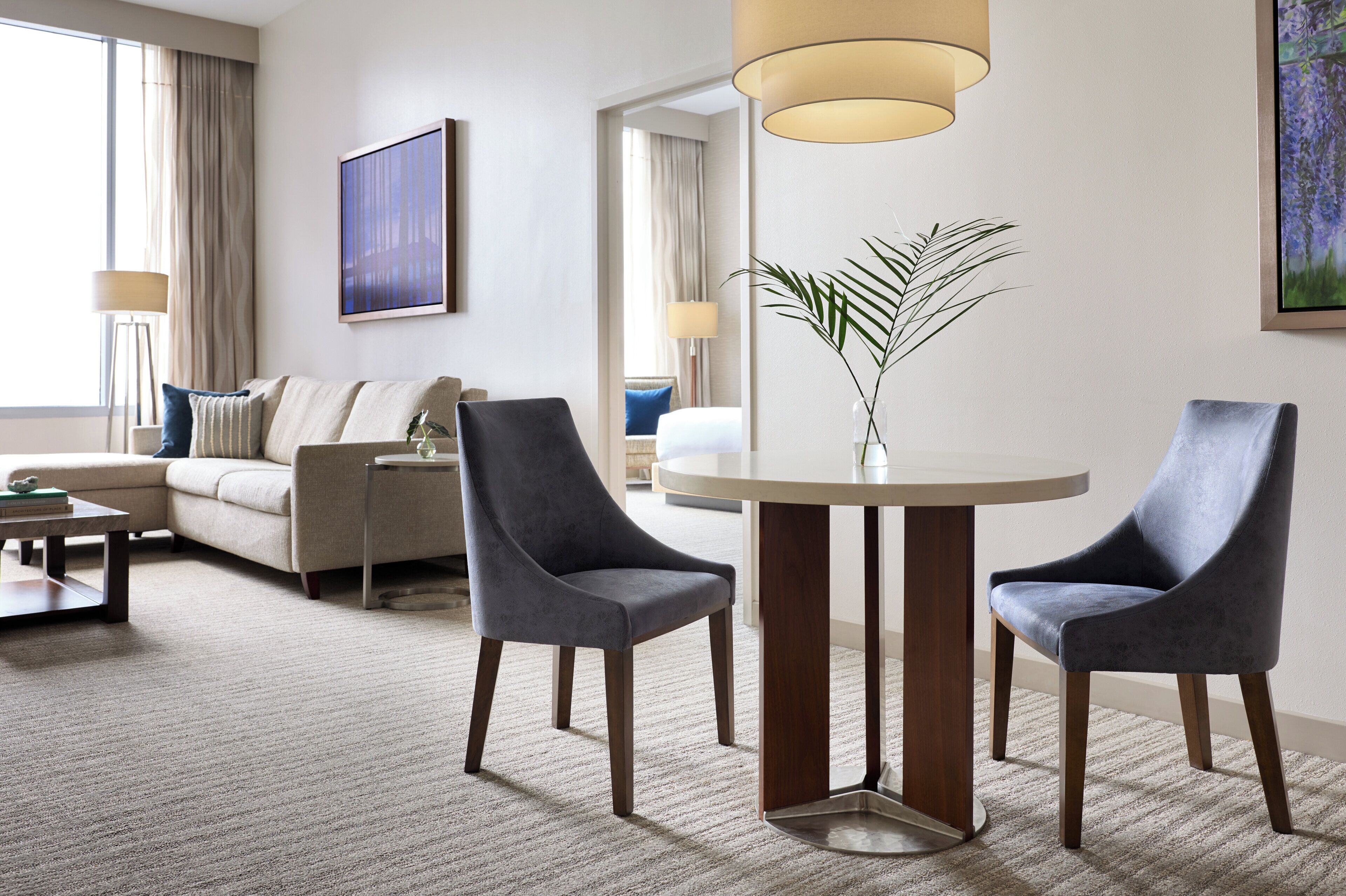 A modern hotel room with a sofa, two armchairs, a round table, and a large window with curtains.
