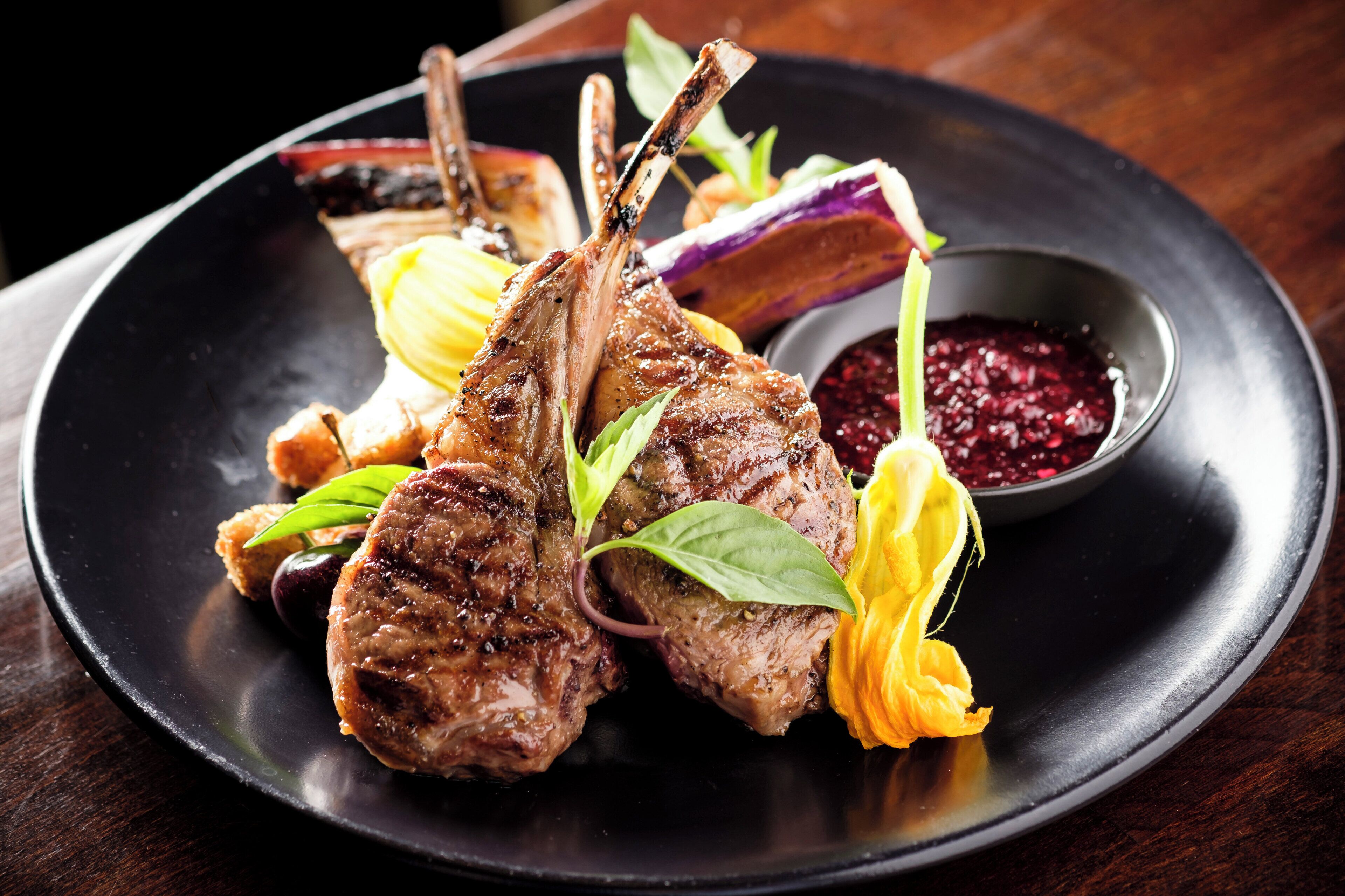Une assiette contenant des côtelettes d’agneau grillées, des légumes et une sauce aux fruits rouges.
