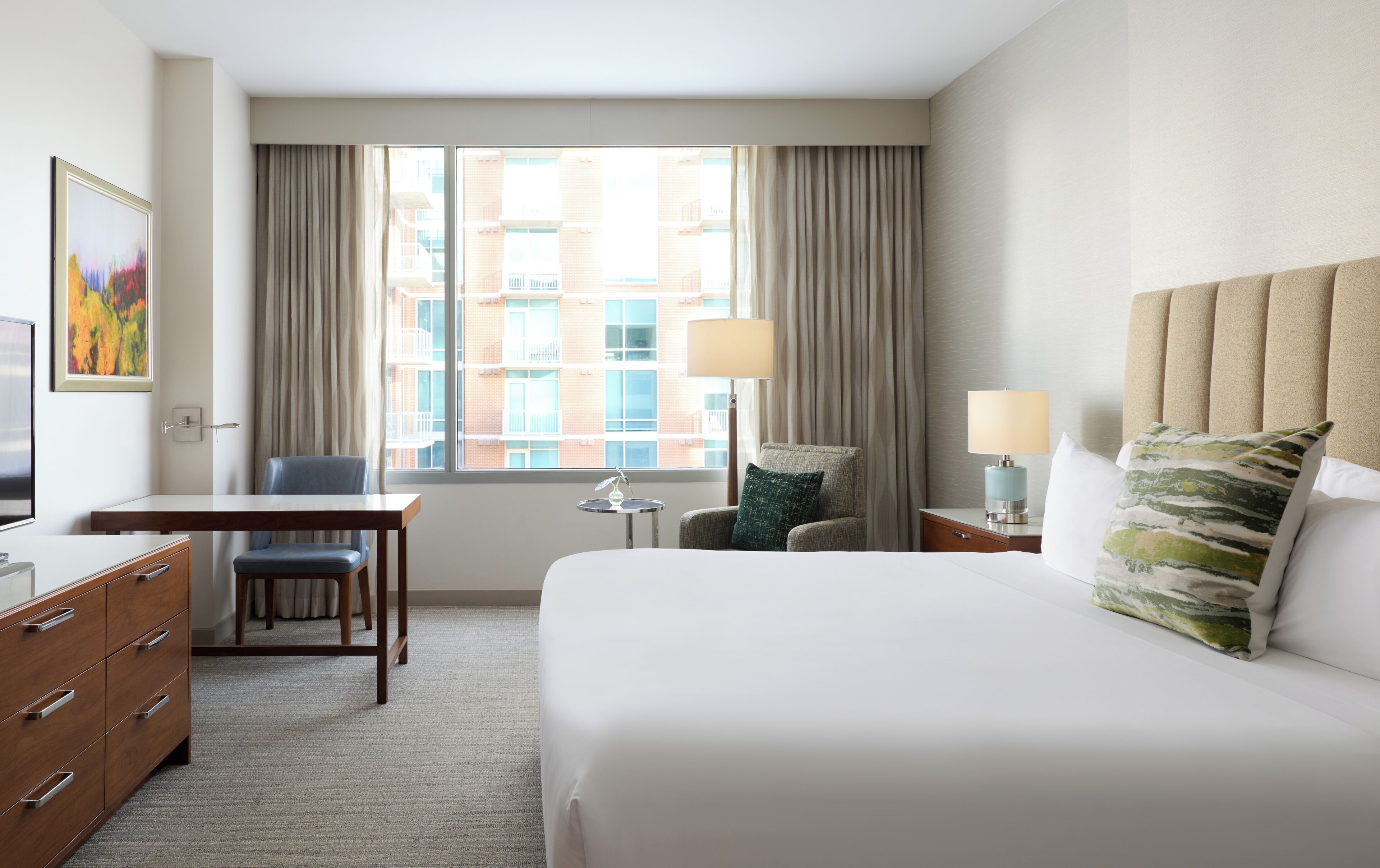 A hotel room with a bed, a desk, a chair, a lamp, and a view of buildings through the window.