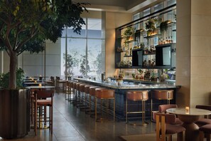 Zona de bar con una barra alargada, varios taburetes y una gran planta en maceta.