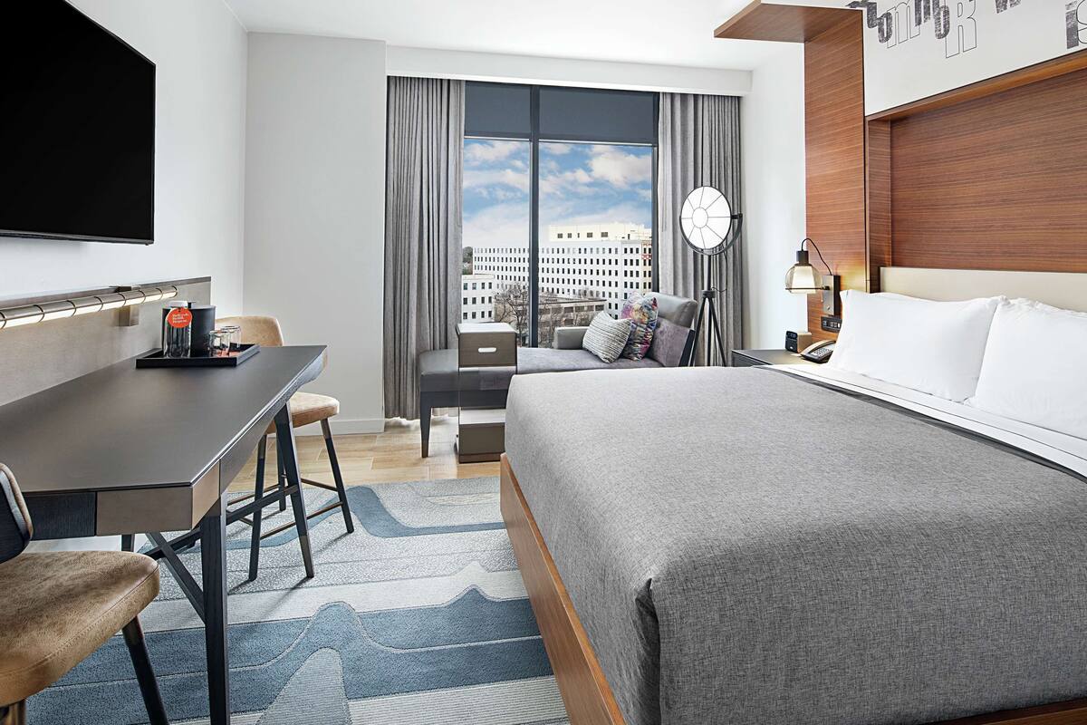 A modern hotel room with a large bed, a desk with a chair, a flat-screen TV, and a view of the city through large windows.