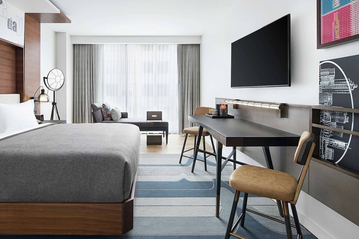 A hotel room with a large bed, a desk, a chair, a TV, and a window with curtains.