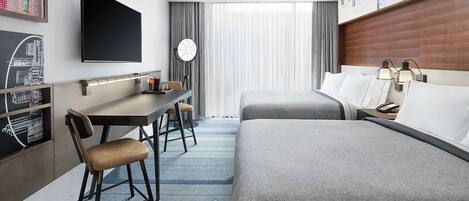 Ein Hotelzimmer mit einem Bett, einem Schreibtisch mit Stuhl, einem Fernseher, einer Uhr und einem Fenster mit Vorhängen.