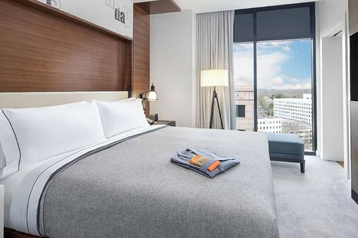 A hotel room with a large bed, a bedside table, a lamp, a TV, and a view of the city.