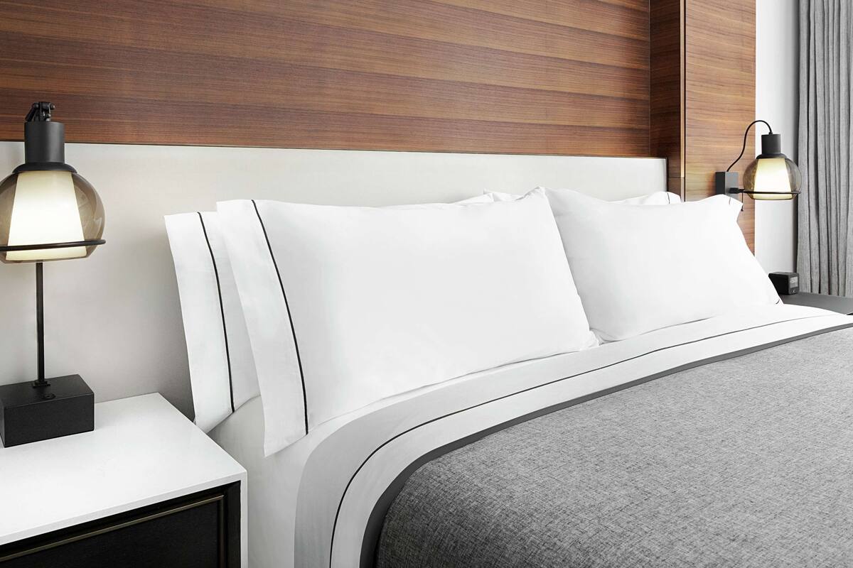 A modern hotel room with a neatly made bed, two bedside lamps, a nightstand, and a wooden headboard.