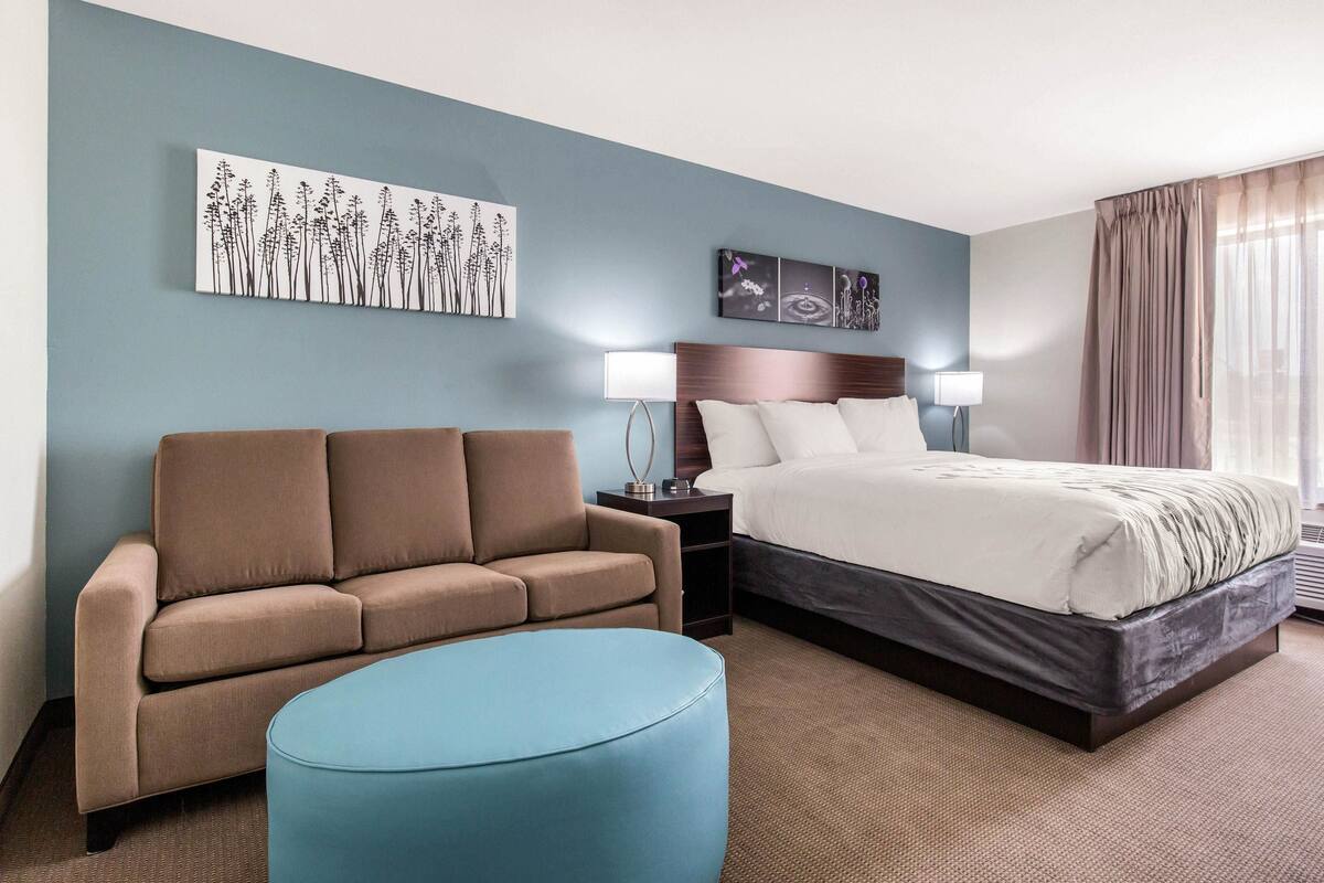 A hotel room with a bed, a sofa, a blue ottoman, a nightstand with a lamp, and wall art.