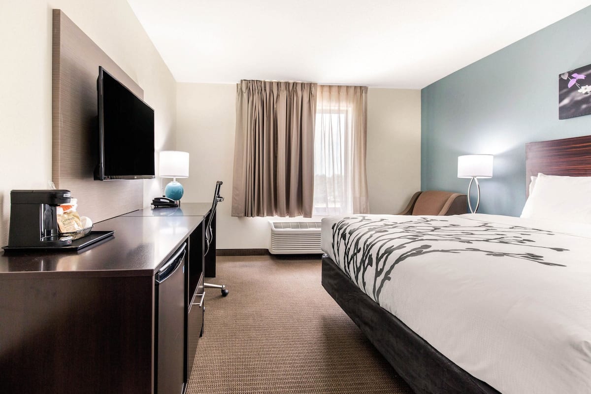 A hotel room with a bed, a desk with a coffee maker, a TV, a window with curtains, and a wall-mounted lamp.