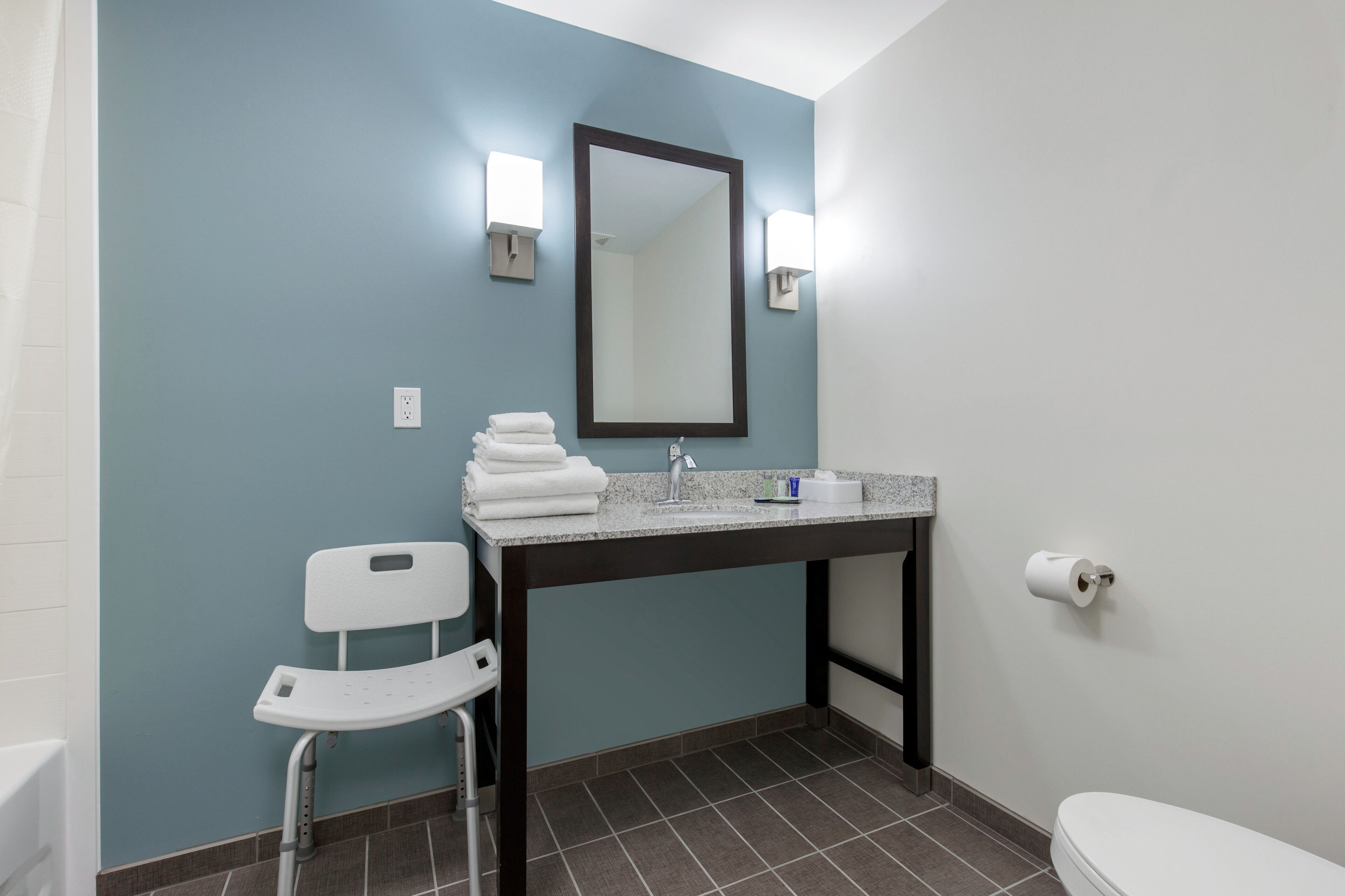 Ein Badezimmer mit blauer Wand, einer weißen Toilette, einem Waschbecken mit Spiegel und einem weißen Stuhl.