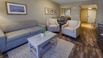 Una sala de estar con un sofá gris, dos sillones, una mesa de centro y una alfombra. Hay cuadros enmarcados en la pared y una cocina al fondo.