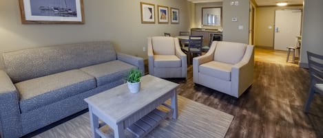 Una sala de estar con un sofá gris, dos sillones, una mesa de centro y una alfombra. Hay cuadros enmarcados en la pared y una cocina al fondo.