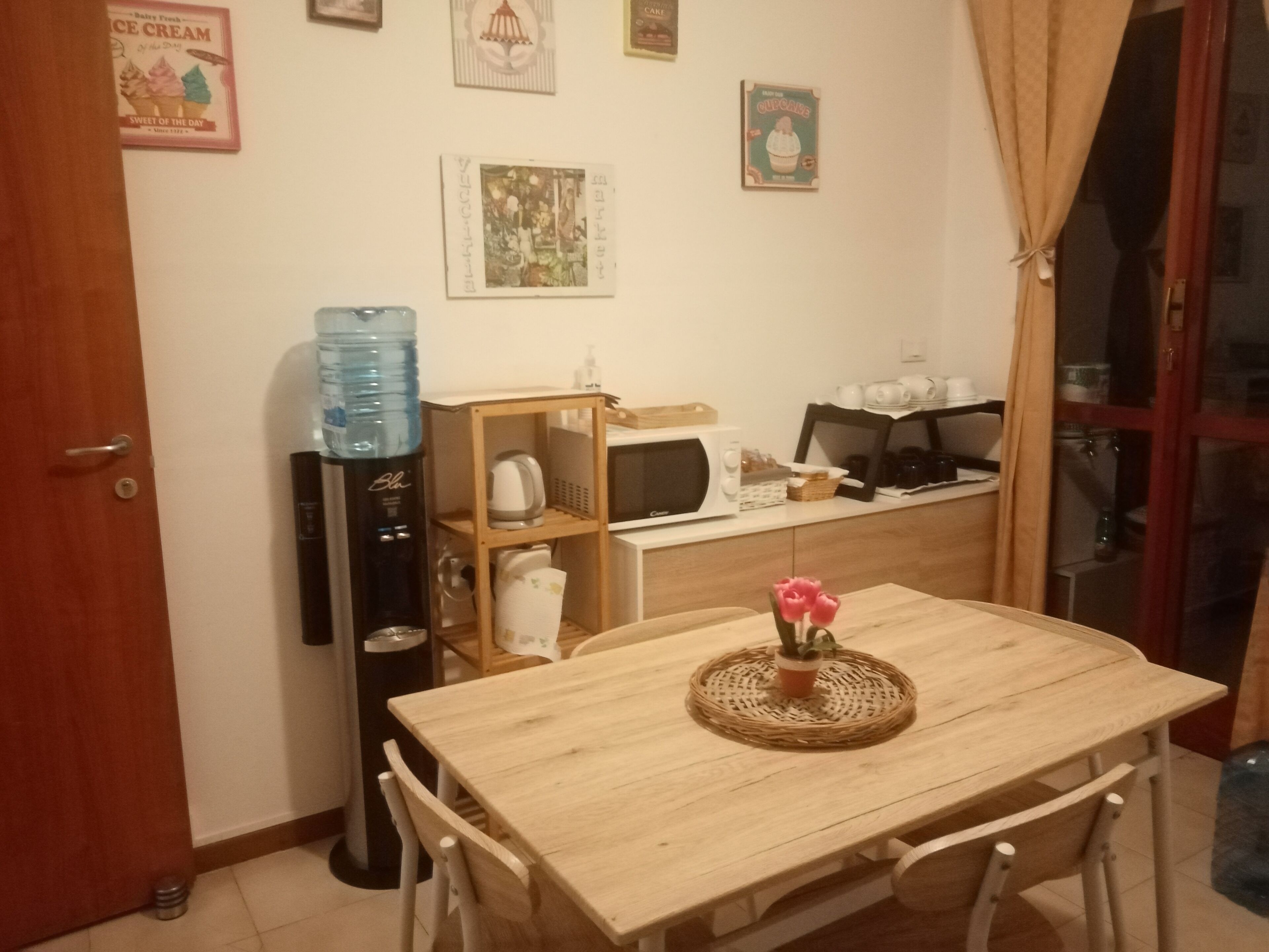 A dining area with a wooden table, chairs, a water dispenser, a microwave, and a shelf with kitchen items.