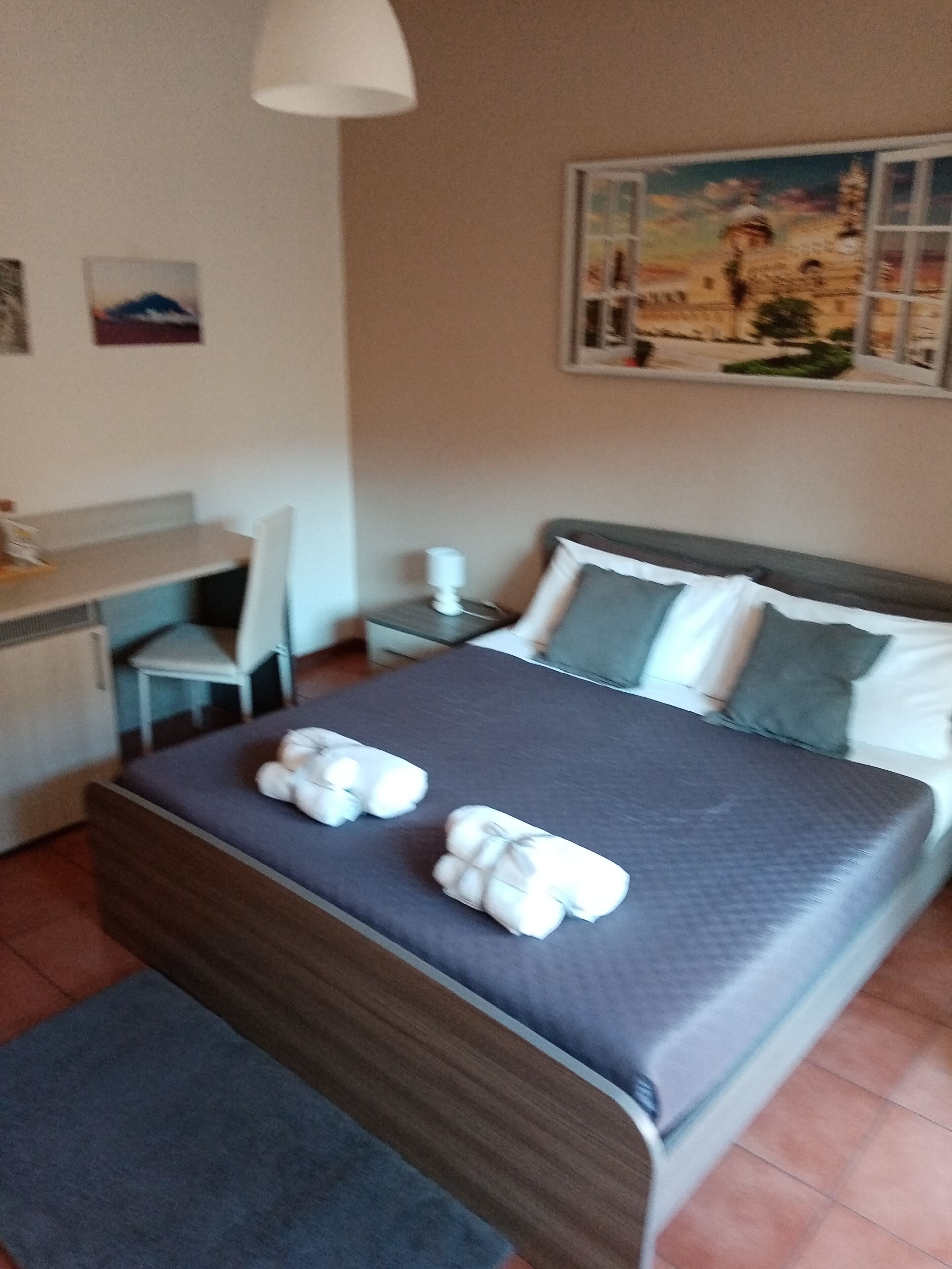 Habitación de hotel ordenada con una cama, un escritorio y una silla. Hay toallas sobre la cama y un cuadro en la pared.