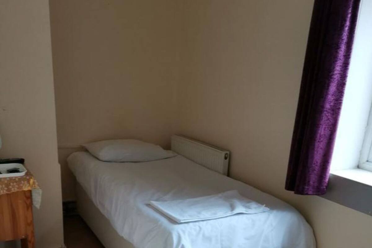 A small, single-bed room with a wooden floor, a radiator, a small table with a basket, and a window with purple curtains.