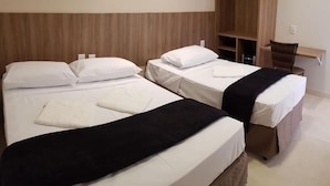 Habitación de hotel con tres camas dispuestas en fila, cabecera de madera, un escritorio pequeño con una silla y un estante en la pared.