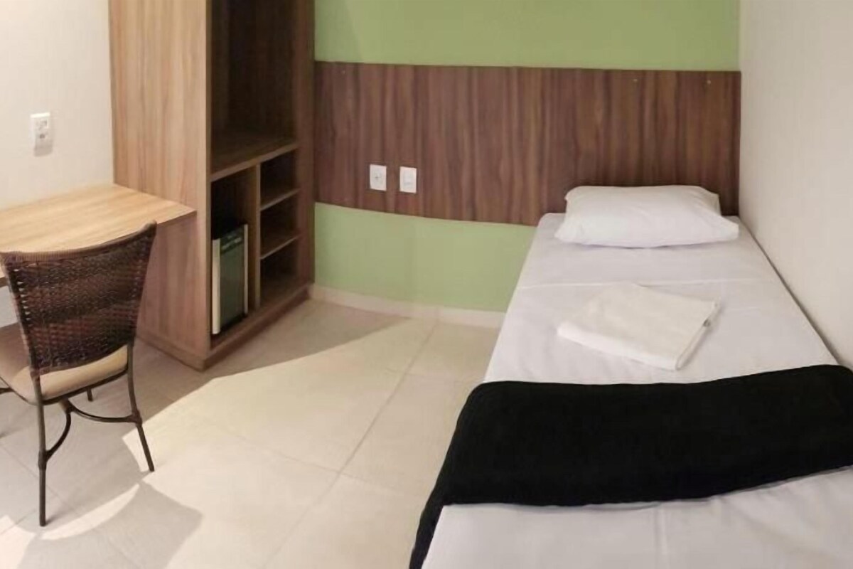 A single bed with a black blanket, a wooden desk with a chair, and a wardrobe in a room with green walls.