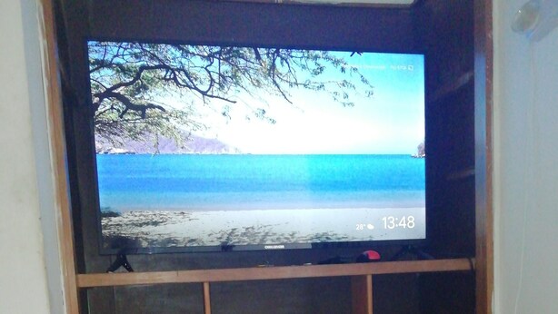 A television on a wooden stand with a beach scene on the screen, a shelf with rolled-up towels, and a tiled floor. - Taminaka Hostel (Santa Marta)