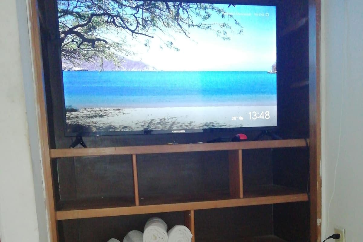 A television on a wooden stand with a beach scene on the screen, a shelf with rolled-up towels, and a tiled floor.