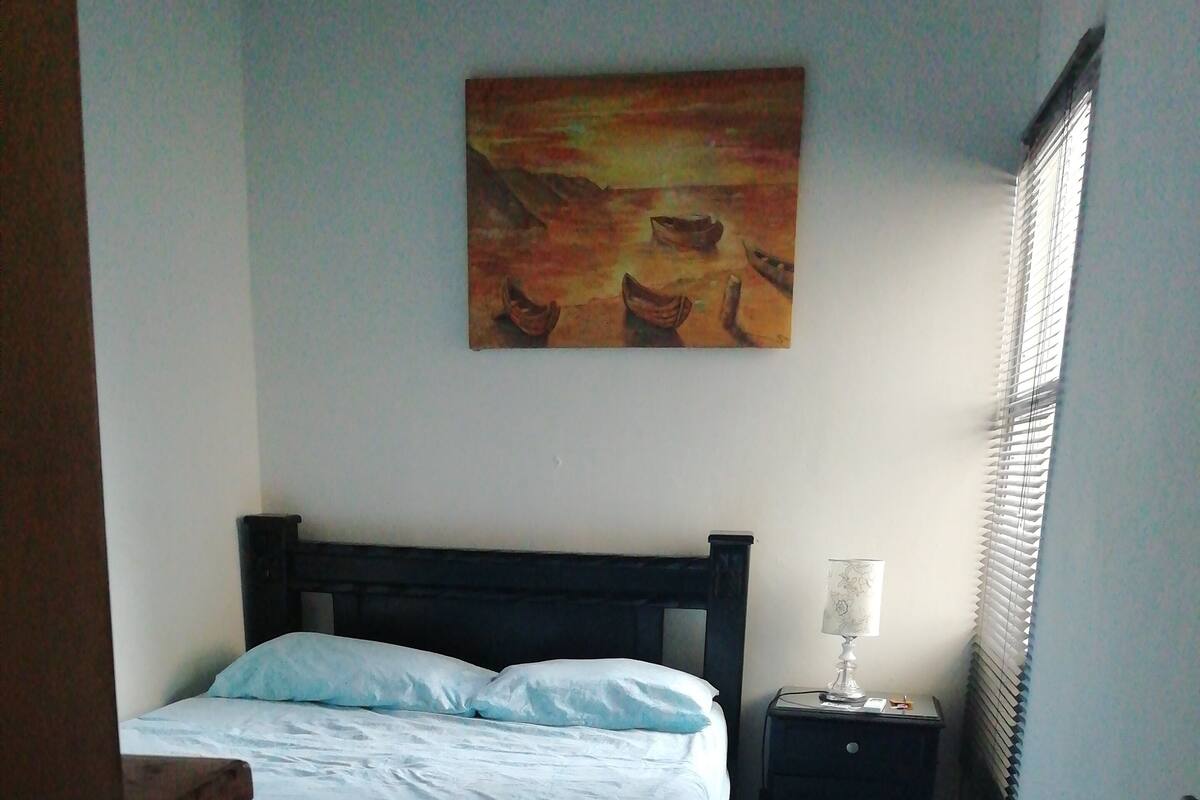 A bedroom with a bed, a nightstand, a lamp, a painting on the wall, and a window with blinds.