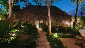 A thatched-roof structure with a stone pathway leading to a door, surrounded by tropical plants and a bench. - Colonte Hotel Origen (Valladolid)