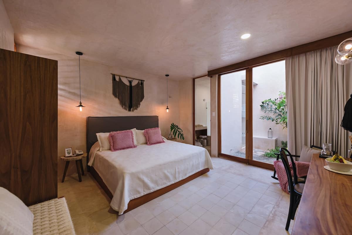 Un dormitorio con una cama grande, mesitas de noche, un armario de madera, una pequeña mesa redonda con una lámpara y vistas al exterior a través de una puerta corredera.