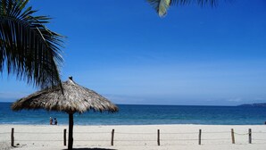 Ein Strand mit einem Reetdachsonnenschirm, Palmen und blauem Himmel.