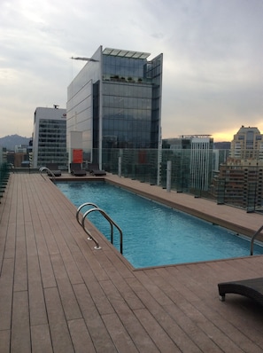 A rooftop pool with a wooden deck, glass railings, and a view of modern buildings. - Rukaza El Golf (Santiago)